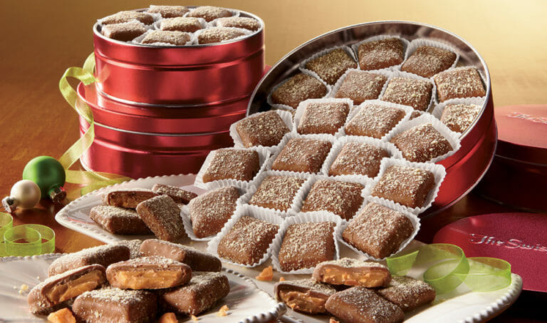 Three sizes of red tins of butter toffee, with a tipped tin allowing some toffee to flow onto decorative plates.